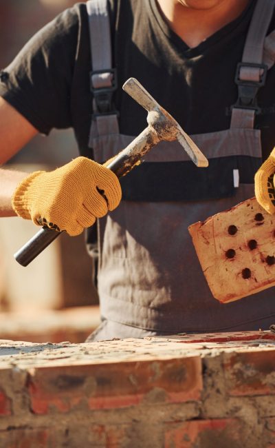 Holding brick and using hammer. Construction worker in uniform and safety equipment have job on building