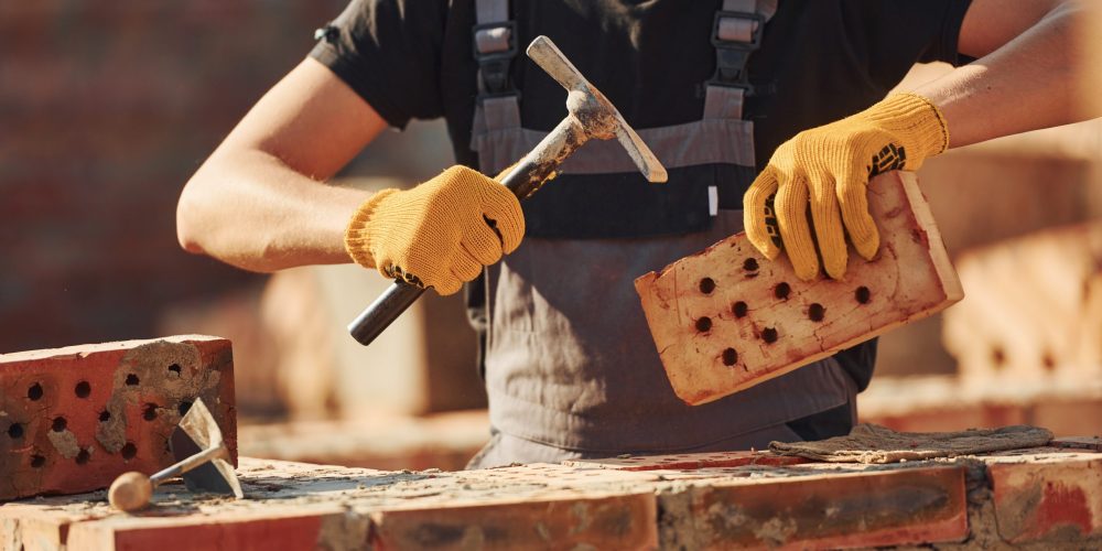 Holding brick and using hammer. Construction worker in uniform and safety equipment have job on building
